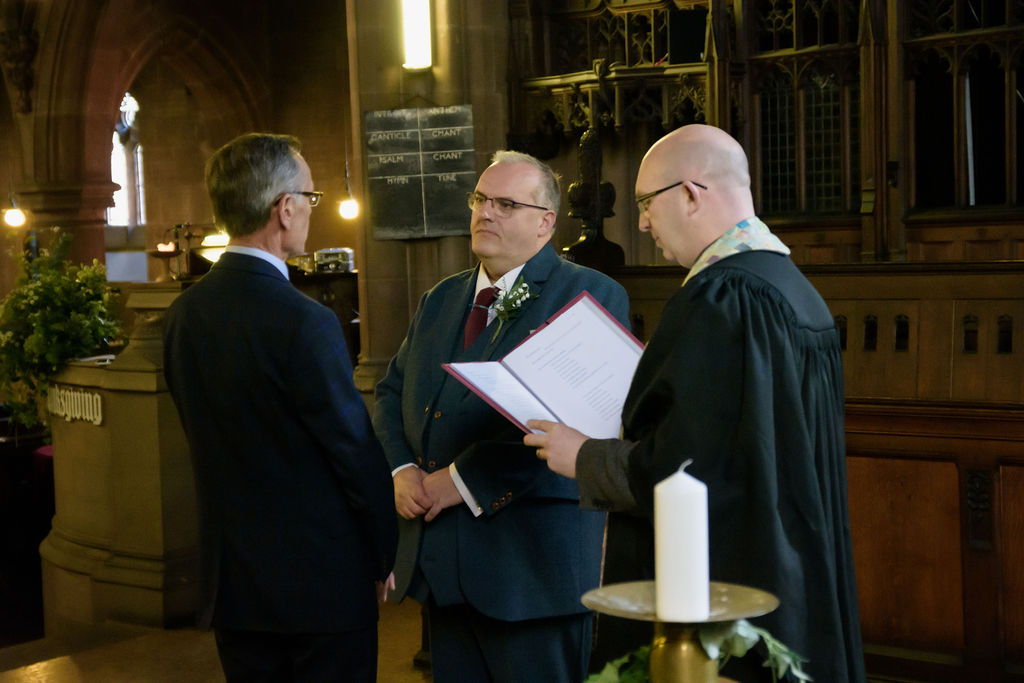 Two smiling grooms stand at the altar of a beautiful church, facing each other and exchanging vows. Feel the love with Cheshire wedding photographer Michael Pardoe