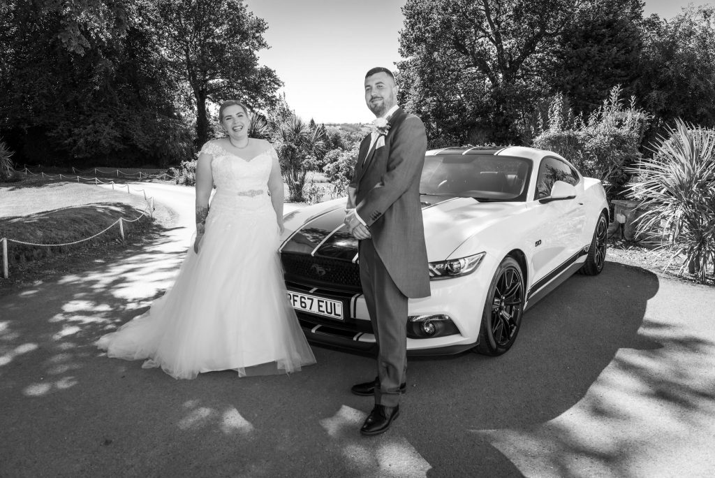 a black and white photograph of a newly married couple stood in front of a Ford Mustang at Highfield Hall during their wedding day