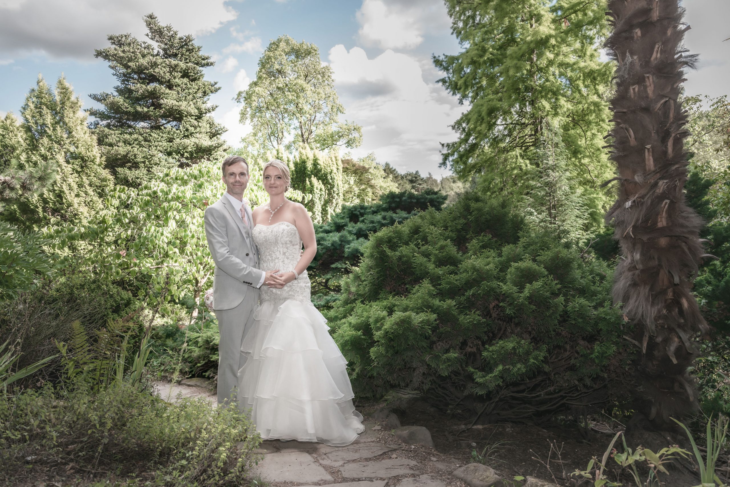 a groom wearing a grey suit holding hands with his bride who is wearing an ivory wedding dress surrounded by trees and greenery in Manchester