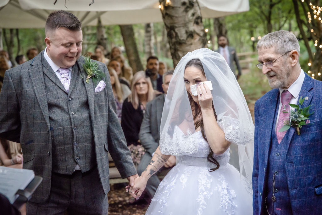 Radiant bride sheds tears of happiness, standing between her proud father and loving groom at their heartfelt wedding ceremony at Cheshire Woodland Weddings