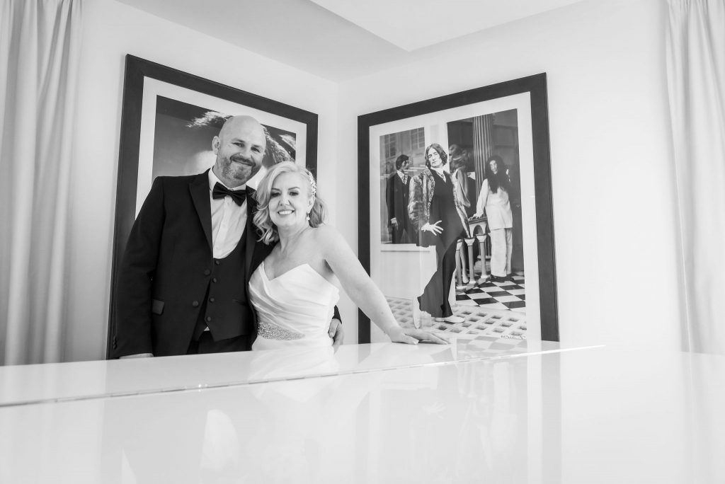 a black and white wedding photograph of a newly married couple stood by a grand piano with a photograph of John Lennon behind them