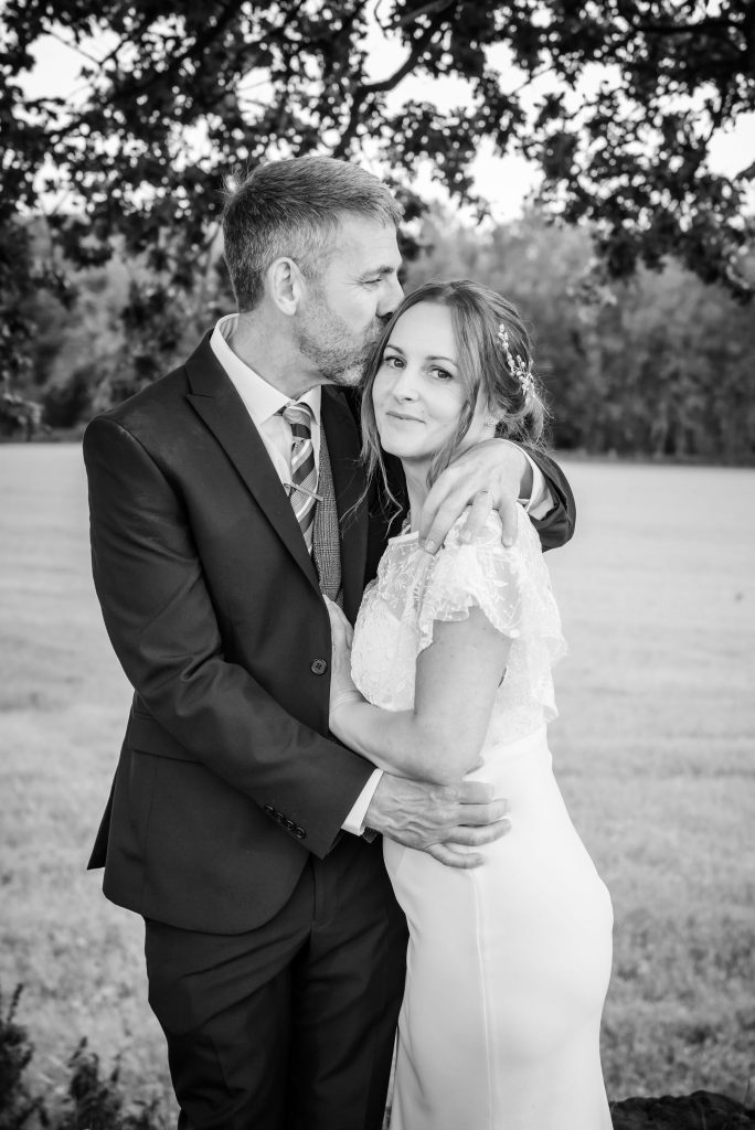 a black and white wedding day photograph of a groom kissing his bride while she smiles