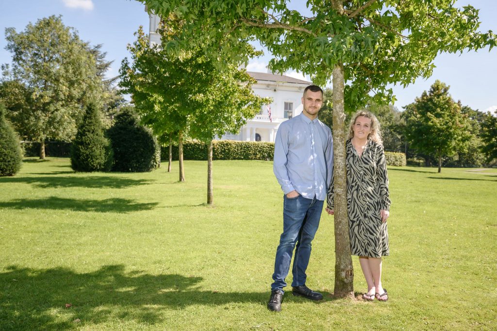 a man wearing a blue shirt and jeans holding hands with his fiance who is wearing a floral patterned dress stood under a tree at Runcorn Town Hall during their pre wedding photo shoot