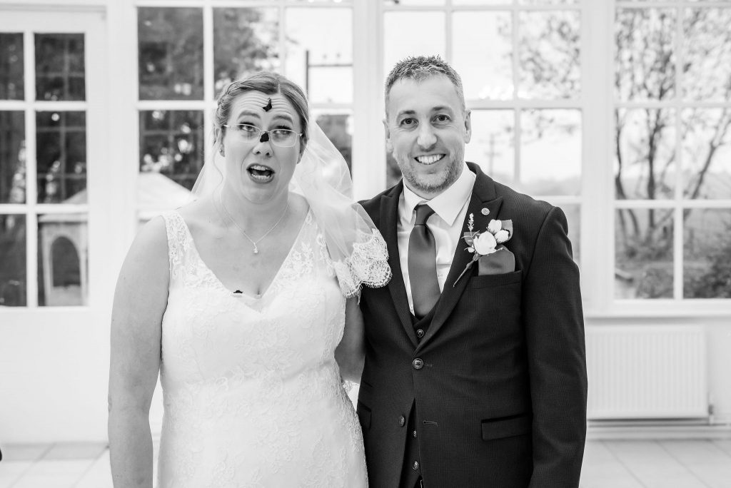 a surprised looking bride blowing confetti off her face stood next to her groom who is smiling at Highfield Hall